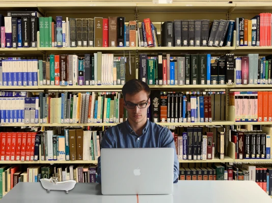 Giovane che lavora con il portatile. Alle spalle, una grande scaffalatura piena di libri. Se sei un editore e hai bisogno di risorse esterne, possiamo offrirti una gamma di servizi di alta qualità