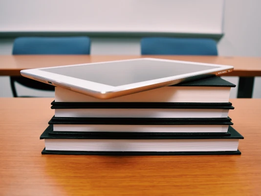Realizziamo la manualistica che ti occorre, sia per la carta sia per il digitale Quattro libri rilegati posati su un banco scolastico. Sopra di essi, un tablet. Realizziamo la manualistica che ti occorre, sia per la carta sia per il digitale